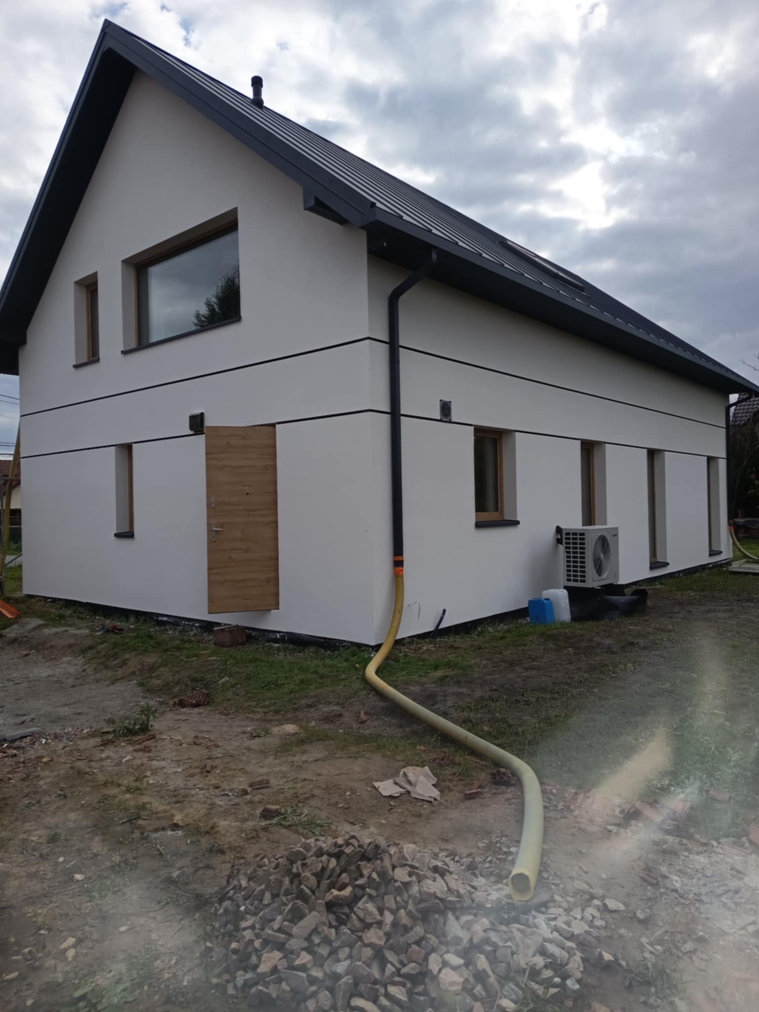 Nowoczesny dom z białą elewacją, ciemnym dachem i drewnianymi oknami. Widoczny fragment ogrodu z kamieniami i wężem. Ujęcie z boku, przy pochmurnym niebie.