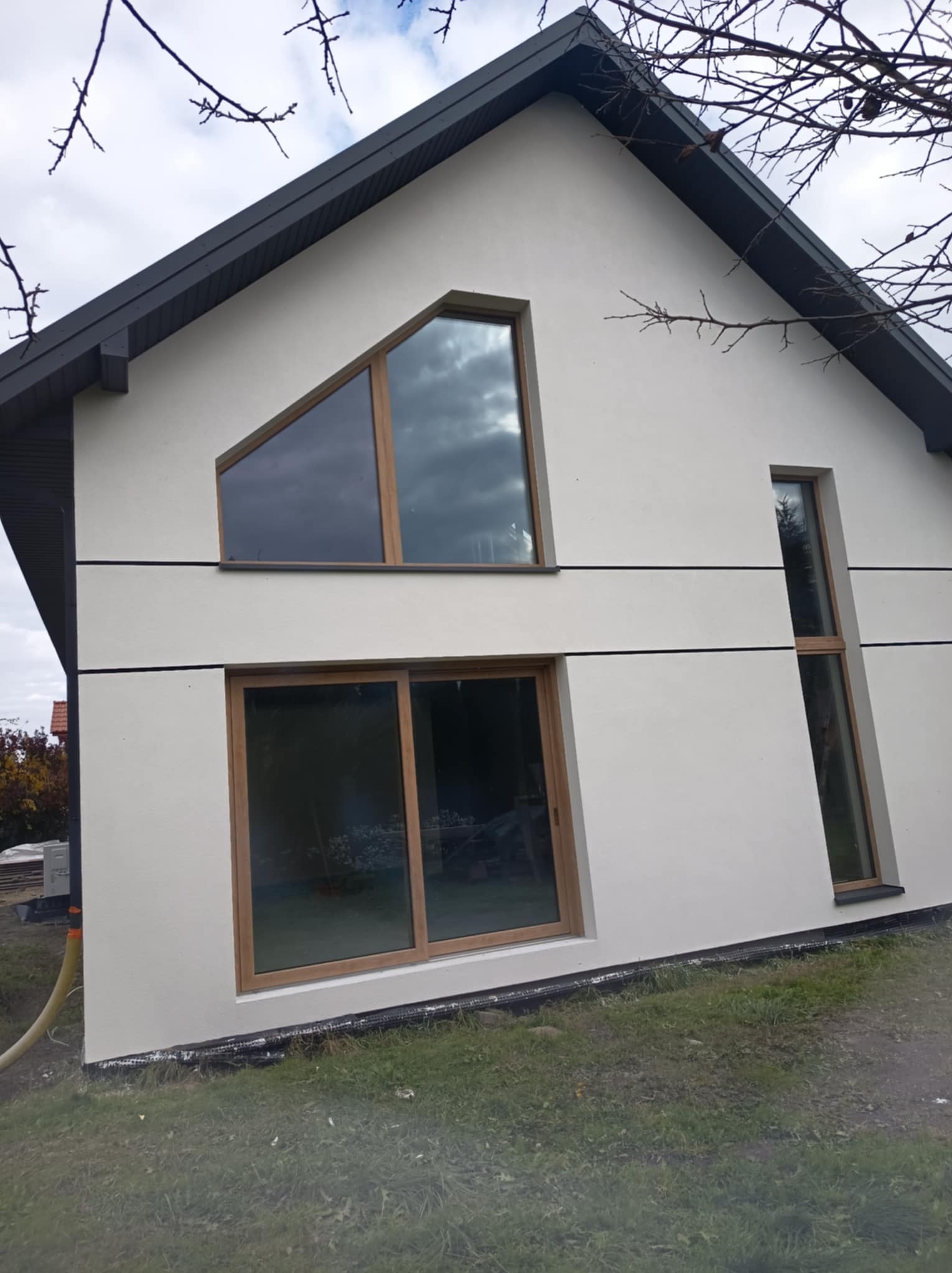 Nowoczesny dom z jasną elewacją, nietypowymi oknami (trójkątne, wąskie), ciemnym dachem i widoczną rurą wentylacyjną przy ścianie. Minimalistyczny design.