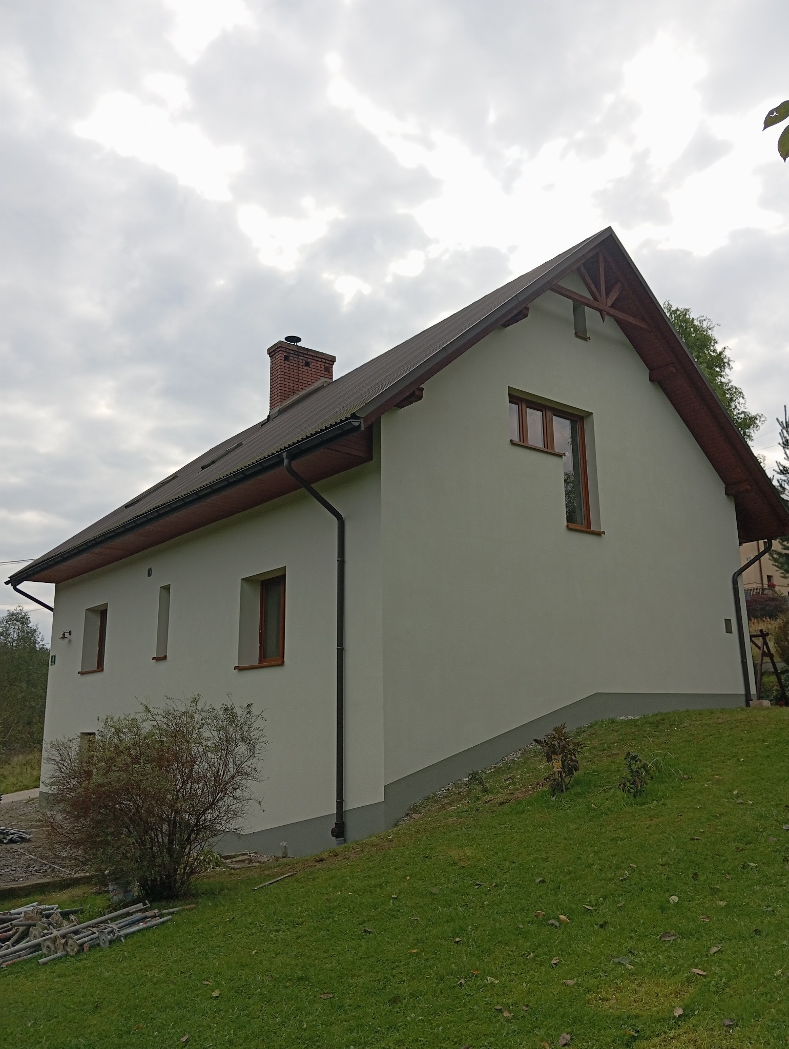 Jasna elewacja domu jednorodzinnego z brązowym dachem i oknami, widok z boku na tle pochmurnego nieba. Na trawniku widoczne elementy rusztowania.