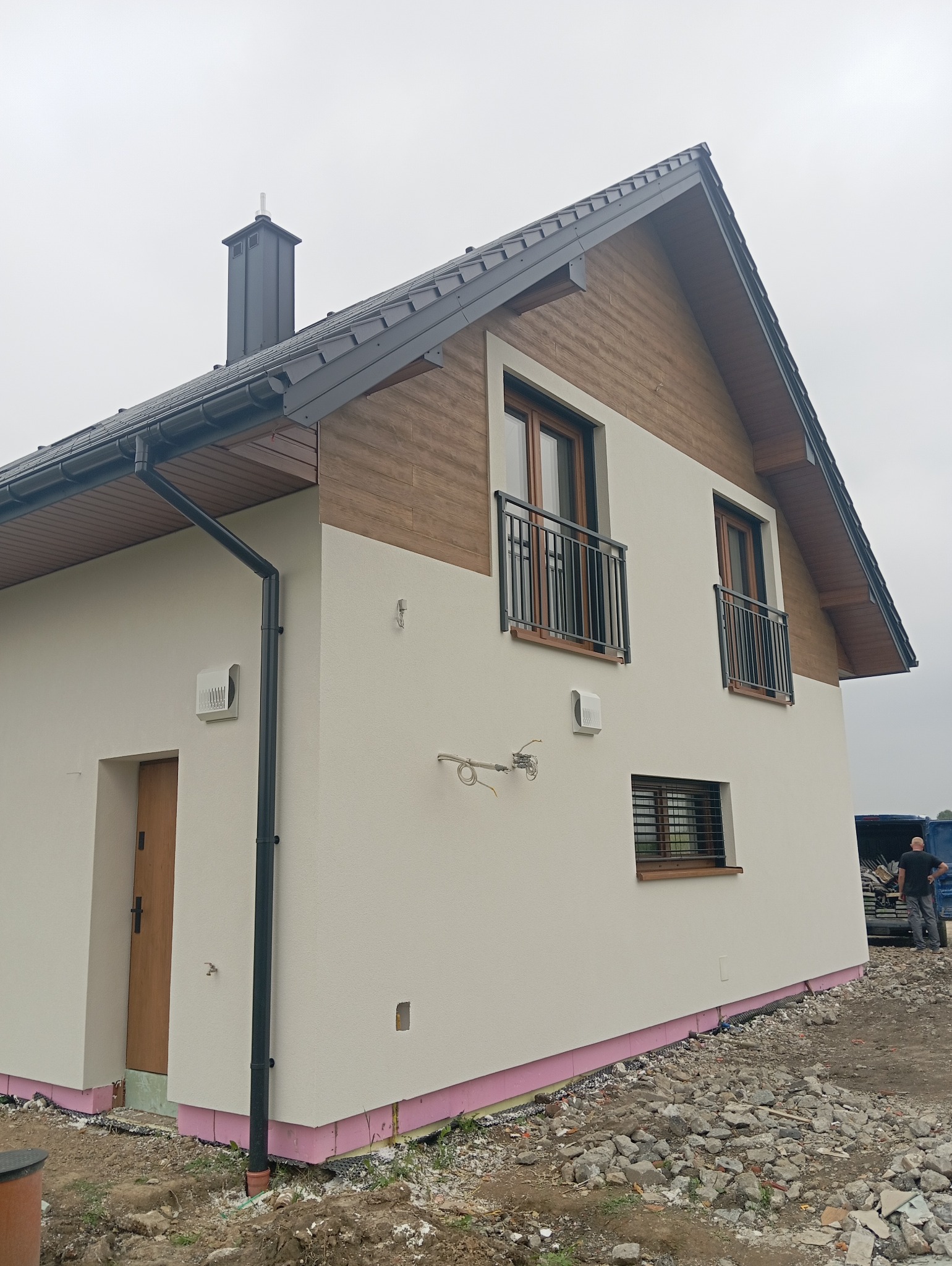 Nowoczesny dom jednorodzinny z elewacją w odcieniach beżu i brązu, z widocznymi elementami wykończeniowymi i fragmentem terenu wokół budynku.