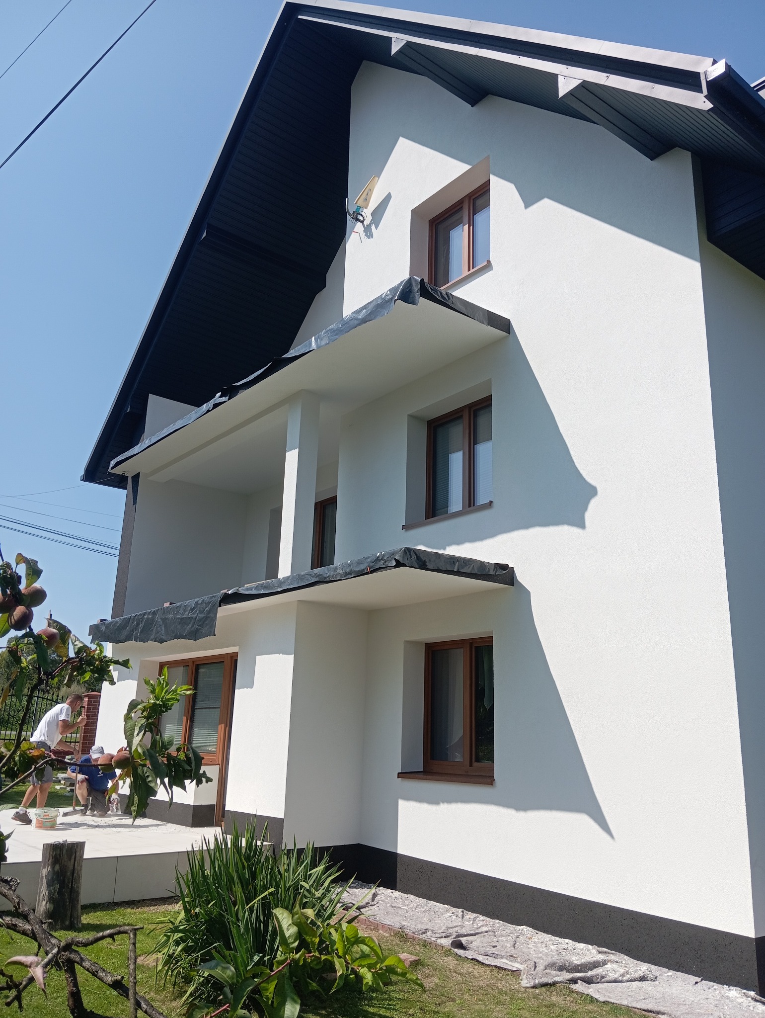 Biała elewacja domu z ciemnym dachem i brązowymi oknami. Na pierwszym planie pracownicy podczas malowania dolnej części elewacji. Widoczne zabezpieczenia.