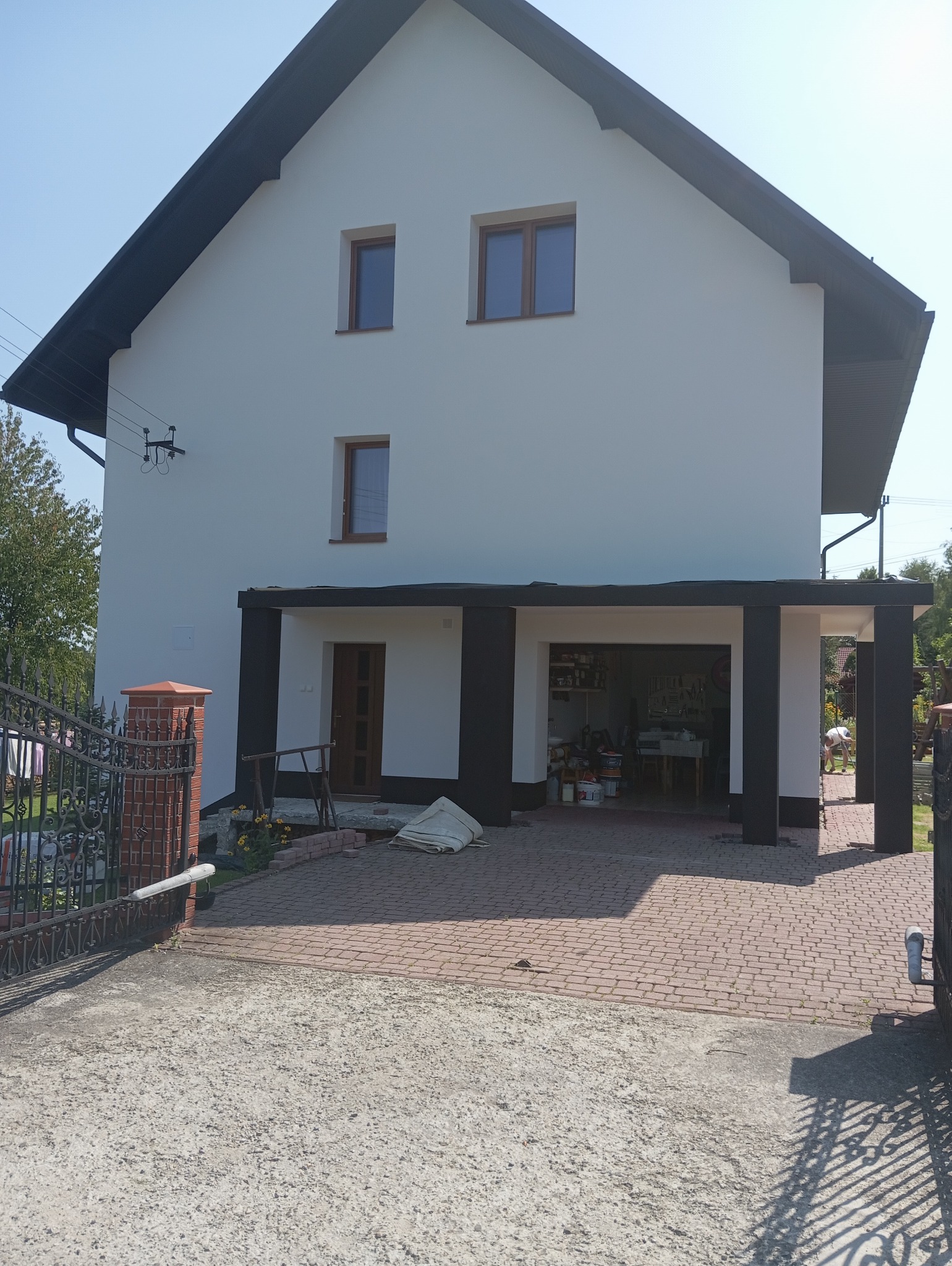 Biały dom z czarnym dachem i kolumnami, widok z zewnątrz. Brama z kutego żelaza na pierwszym planie. Widoczne narzędzia i materiały budowlane w garażu.