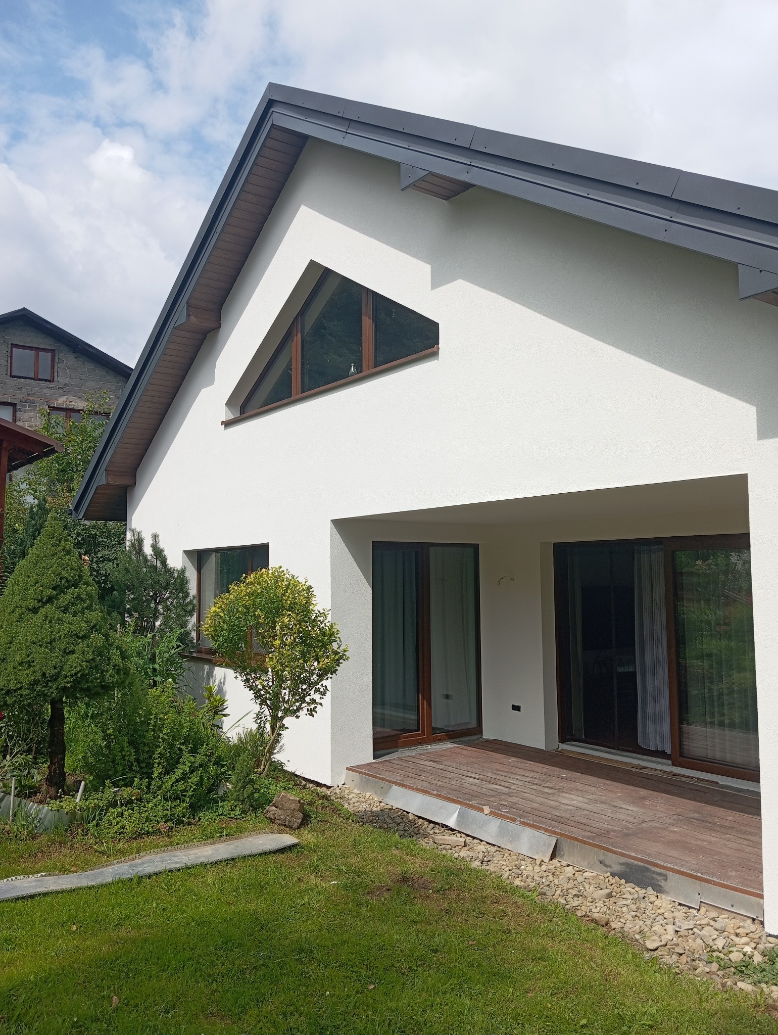 Nowoczesny dom z białą elewacją, drewnianymi oknami i szarym dachem. Widoczne elementy architektury, takie jak trójkątne okno i drewniany taras.