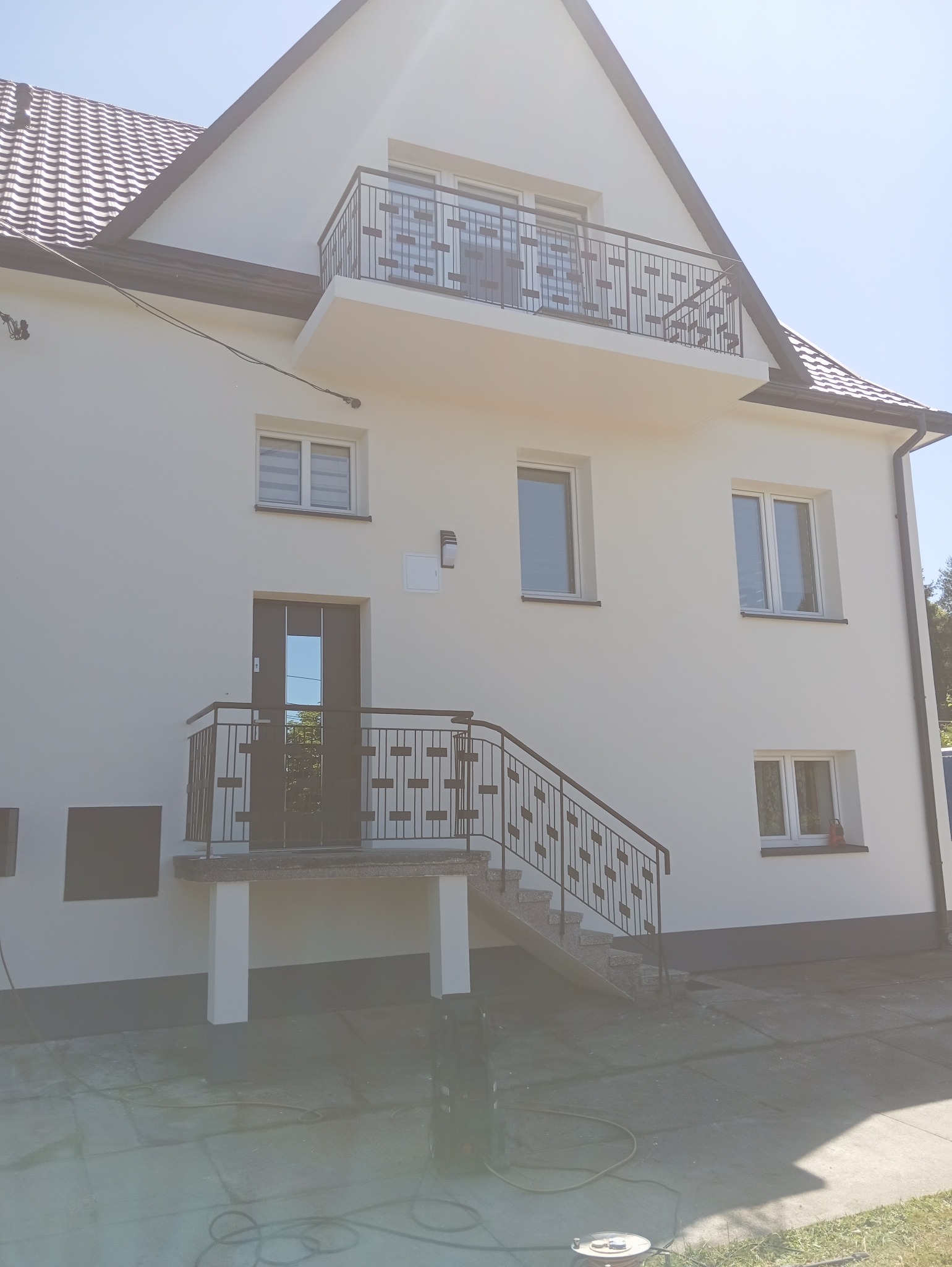 Biały dom jednorodzinny po renowacji elewacji z balkonem i schodami wejściowymi. Widoczne okna, drzwi i elementy wykończeniowe. Na dole wąż i myjka ciśnieniowa.