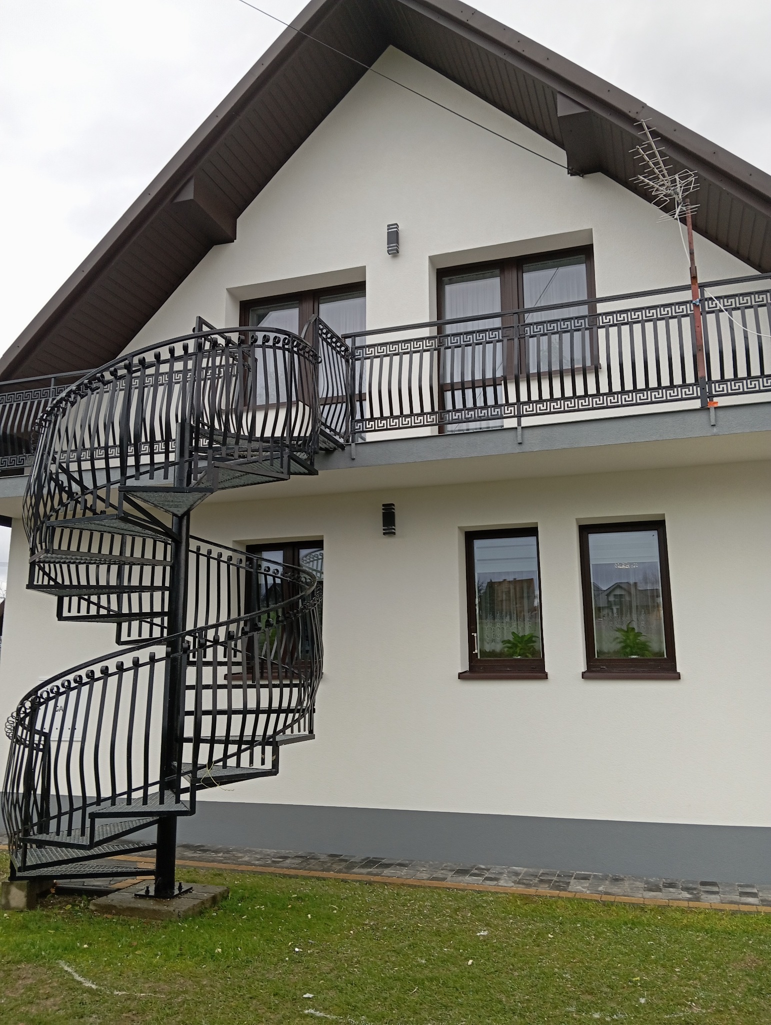 Elewacja domu jednorodzinnego z balkonem i spiralnymi schodami zewnętrznymi. Biała ściana, brązowe okna i dach. Widoczne detale wykończenia.