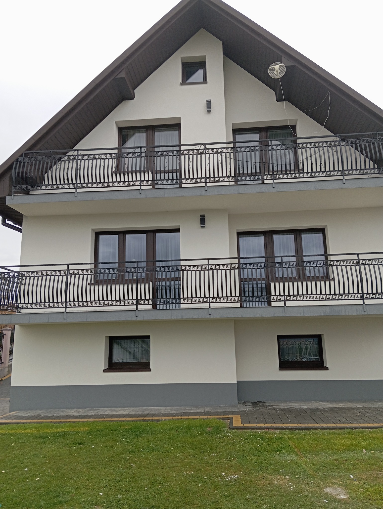 Dwupiętrowy dom z balkonami o ozdobnych, metalowych balustradach w grecki wzór. Świeżo pomalowana elewacja w odcieniach bieli i szarości. Widoczne okna z brązowymi ramami.