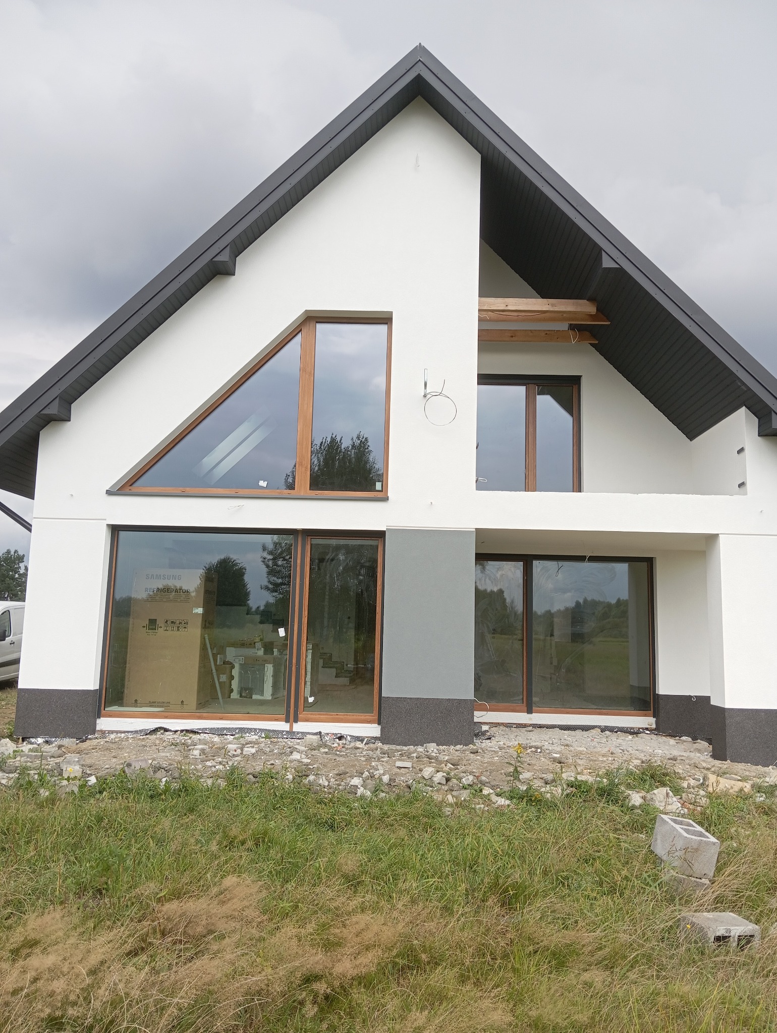 Nowoczesny dom jednorodzinny z białymi ścianami i szarym dachem, duże okna z drewnianymi ramami. Widoczne elementy instalacji na elewacji. Teren wokół domu w trakcie prac wykończeniowych.