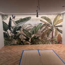 NOW GRES DAMIAN NOWAK - Pokój po remoncie: parkiet w jodełkę, ściana z motywem tropikalnym, listwy LED pod sufitem. Część podłogi zabezpieczona kartonem.