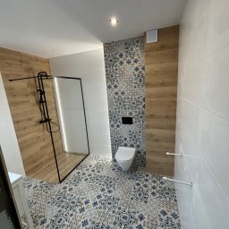 NOW GRES DAMIAN NOWAK - Nowoczesna łazienka po remoncie: kabina prysznicowa z czarną ramą, toaleta, białe płytki ścienne i podłoga w dekoracyjne wzory. Drewnopodobne akcenty na ścianach.