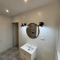 NOW GRES DAMIAN NOWAK - Wykończona łazienka w Szczekocinach: białe płytki na ścianach, wzorzysta podłoga, okrągłe lustro z oświetleniem i biała szafka pod umywalką. Nowoczesny design.