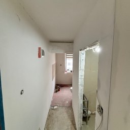 NOW GRES DAMIAN NOWAK - Korytarz w trakcie remontu: ściany po gładziowaniu, widoczne narzędzia, przygotowane otwory na gniazdka, surowa podłoga. Widok na łazienkę w trakcie wykończenia.