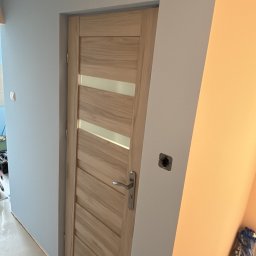NOW GRES DAMIAN NOWAK - Jasne, drewniane drzwi z poziomymi przeszkleniami w świeżo pomalowanym na niebiesko pomieszczeniu. Widoczne narzędzia malarskie i gniazdko elektryczne.