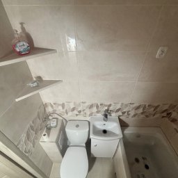 NOW GRES DAMIAN NOWAK - Jasna łazienka po remoncie: beżowe płytki, umywalka z szafką, toaleta i wanna z hydromasażem. Na półkach mydło i drobne akcesoria. Widok z góry.