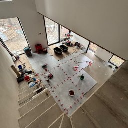 NOW GRES DAMIAN NOWAK - Układanie szarych płytek na podłodze w jasnym wnętrzu, widok z góry na schody i okna. Użyto czerwonych krzyżyków dystansowych. Część podłogi jeszcze nie wykończona.