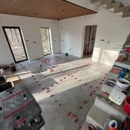 NOW GRES DAMIAN NOWAK - Układanie płytek podłogowych w jasnym wnętrzu z drewnianym sufitem lamelowym. Widoczne narzędzia glazurnicze i krzyżyki dystansowe.