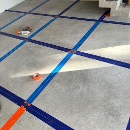 NOW GRES DAMIAN NOWAK - Przygotowanie podłoża pod płytki: wyznaczanie siatki z użyciem taśm i krzyżyków dystansowych na szarym, surowym betonie. Widoczne narzędzia glazurnicze i schody.