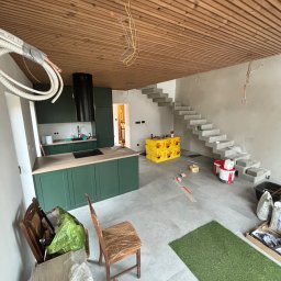 NowGres - Wnętrze w trakcie wykańczania: zielona zabudowa kuchenna, surowe betonowe schody, drewniany sufit lamelowy. Widoczne materiały budowlane i narzędzia.