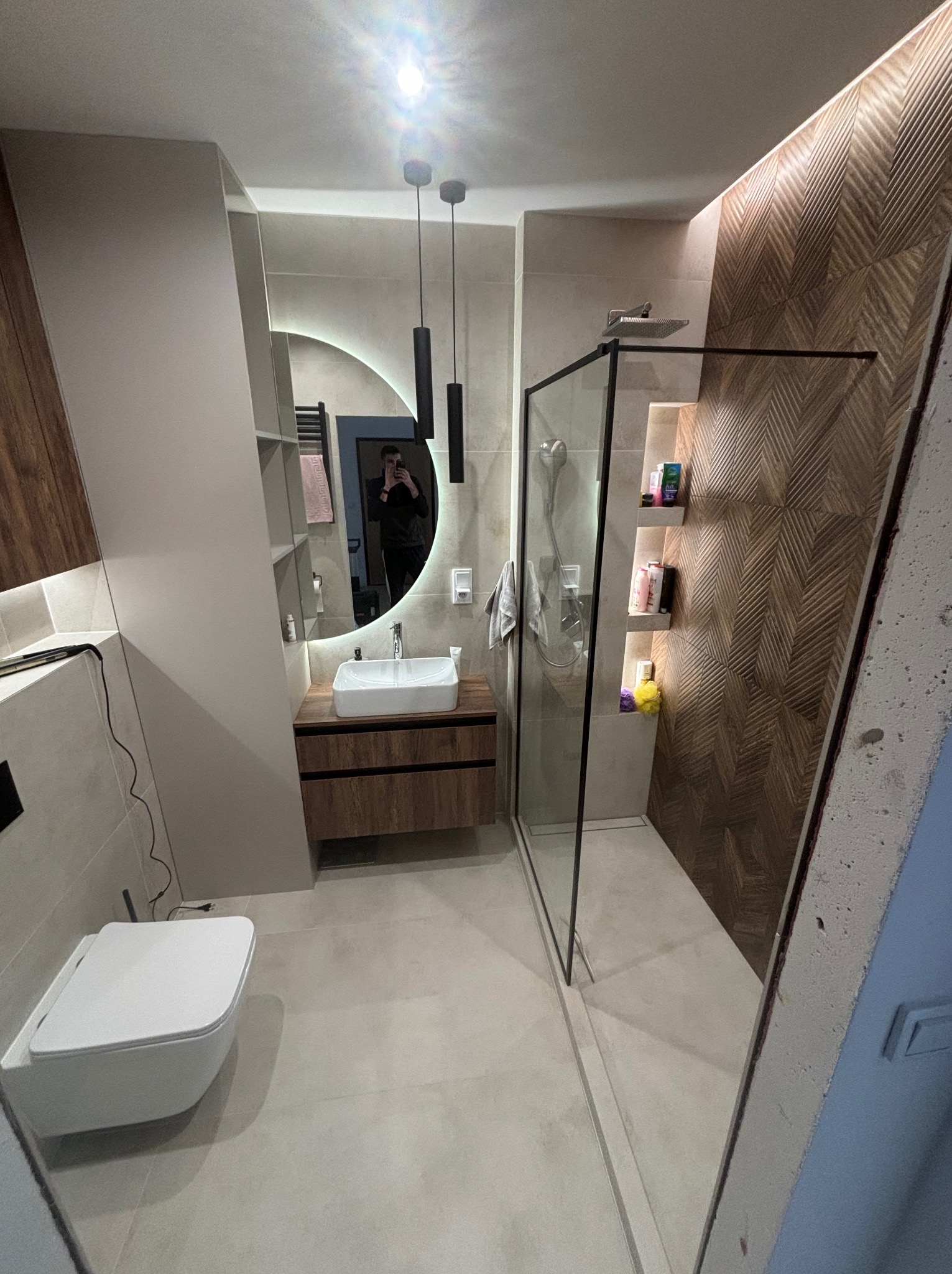 Nowoczesna łazienka po remoncie: kabina prysznicowa typu walk-in, umywalka z szafką z drewnianym fornirem, okrągłe lustro z podświetleniem LED i dekoracyjna ściana z płytek.