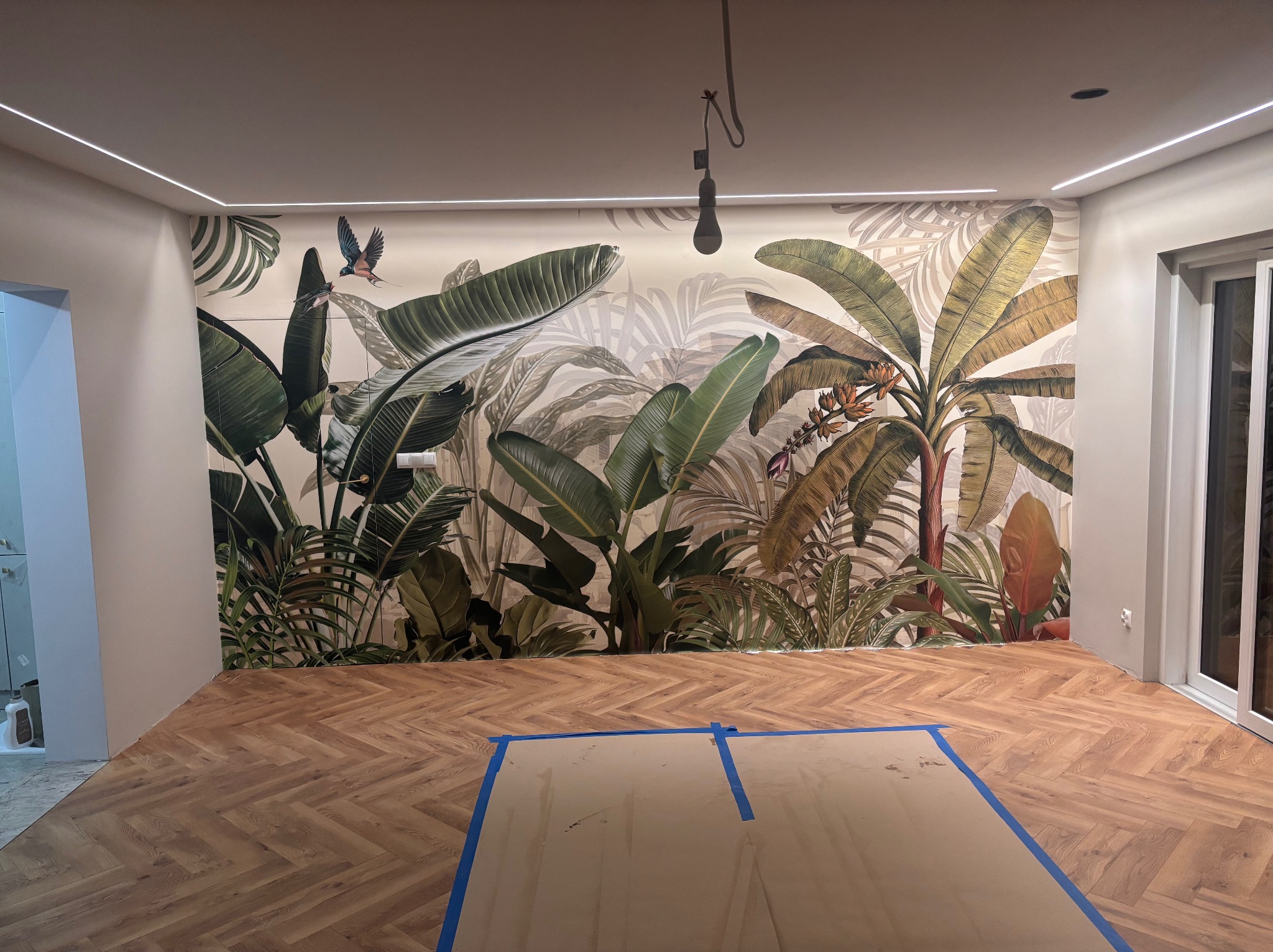 Pokój po remoncie: parkiet w jodełkę, ściana z motywem tropikalnym, listwy LED pod sufitem. Część podłogi zabezpieczona kartonem.