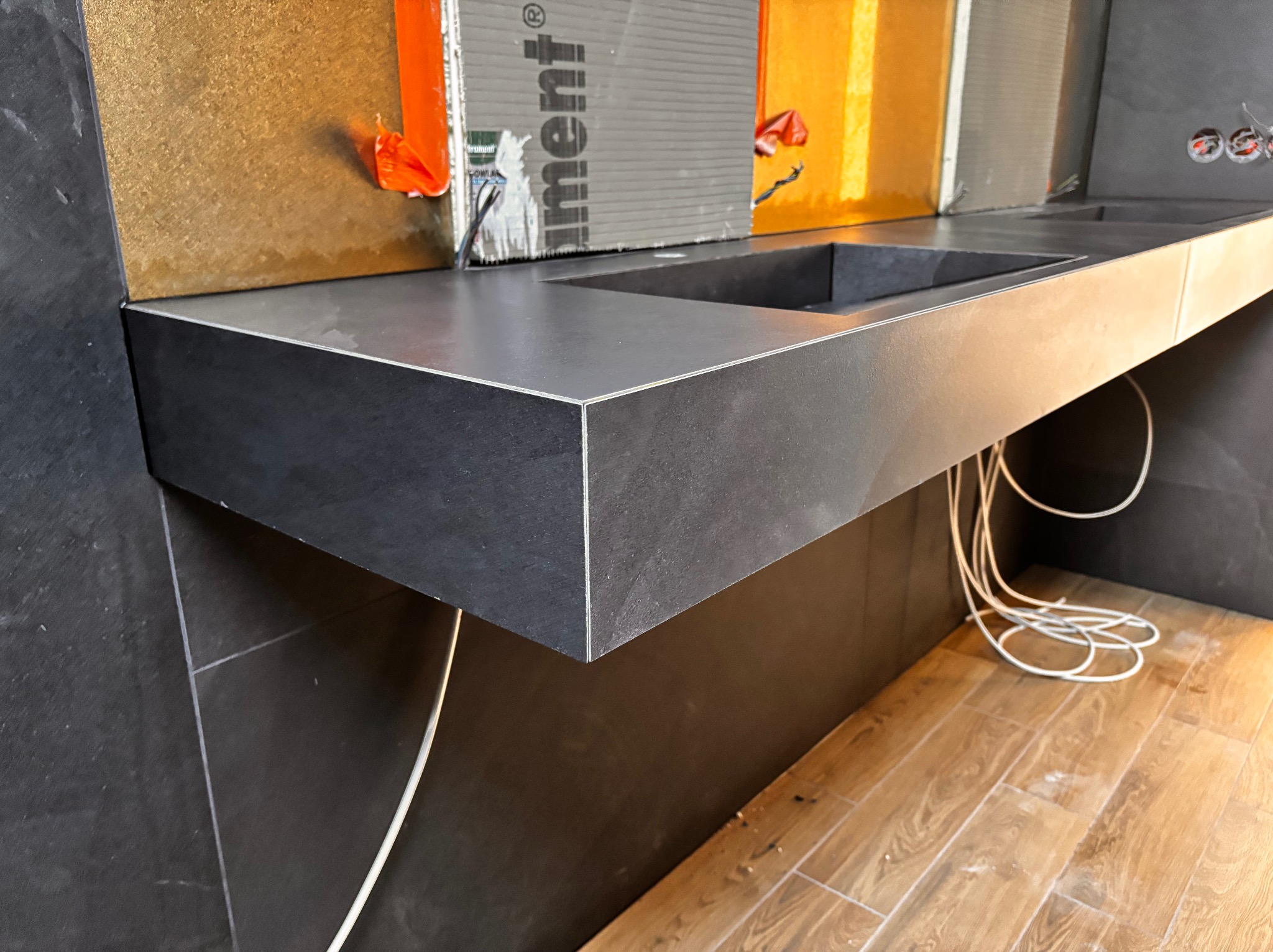 Nowoczesna, czarna umywalka z blatem z płytek w łazience w trakcie wykończenia. Widoczne przewody i fragmenty płytek na ścianie. Podłoga wyłożona panelami.