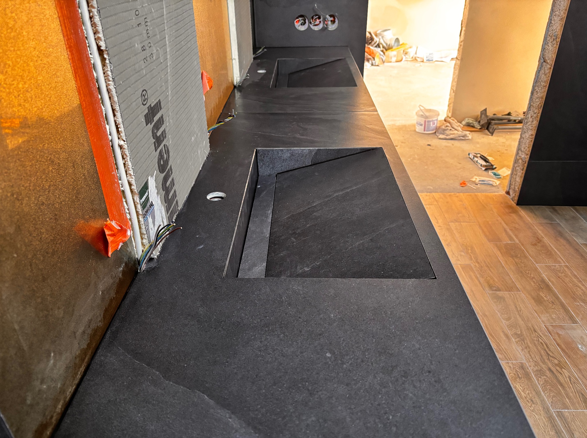 Ciemny blat z dwoma wpuszczonymi umywalkami o kwadratowym kształcie, widoczne przygotowania do montażu płytek na ścianie. W tle remont.