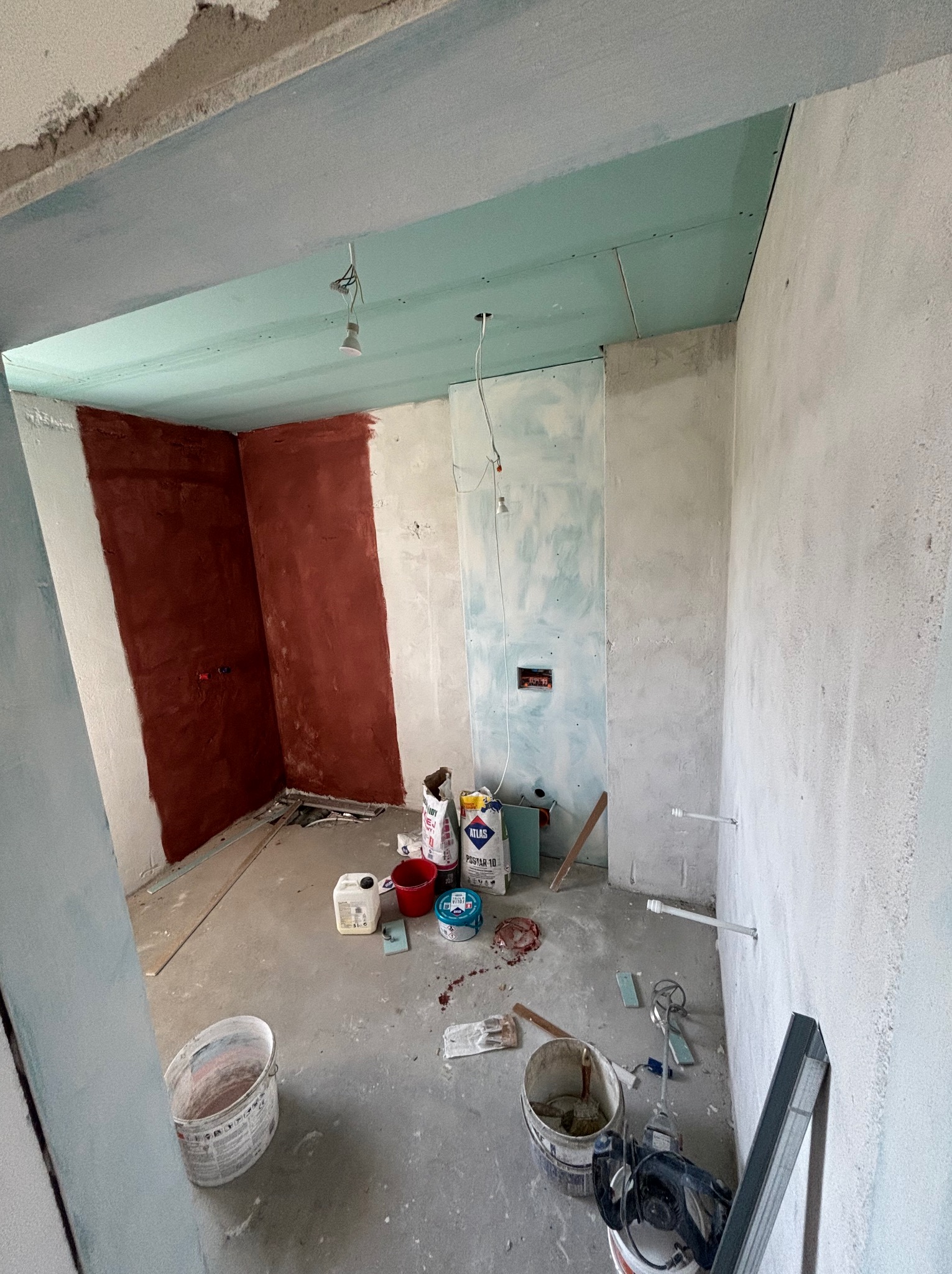 Wnętrze w trakcie remontu: surowe ściany z fragmentami gładzi, widoczne instalacje, worki z zaprawą i narzędzia malarskie. Sufit z płyt kartonowo-gipsowych.