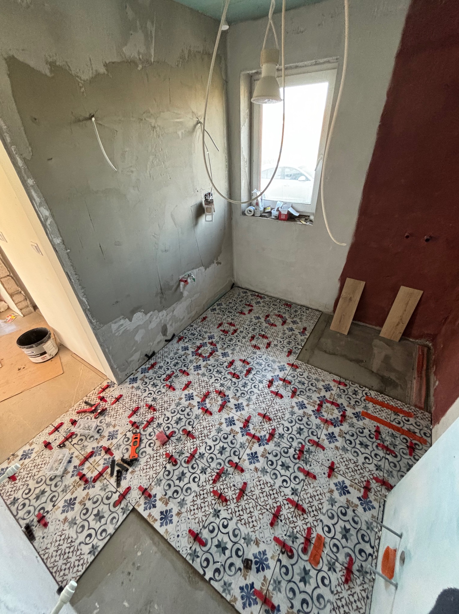 Układanie wzorzystych płytek podłogowych w łazience, z użyciem czerwonych krzyżyków dystansowych. Ściany częściowo przygotowane pod wykończenie, widoczne instalacje.