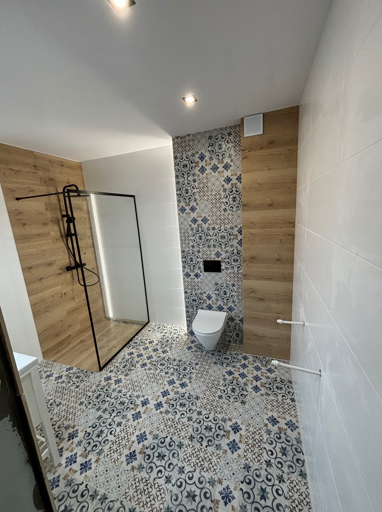 Nowoczesna łazienka po remoncie: kabina prysznicowa z czarną ramą, toaleta, białe płytki ścienne i podłoga w dekoracyjne wzory. Drewnopodobne akcenty na ścianach.
