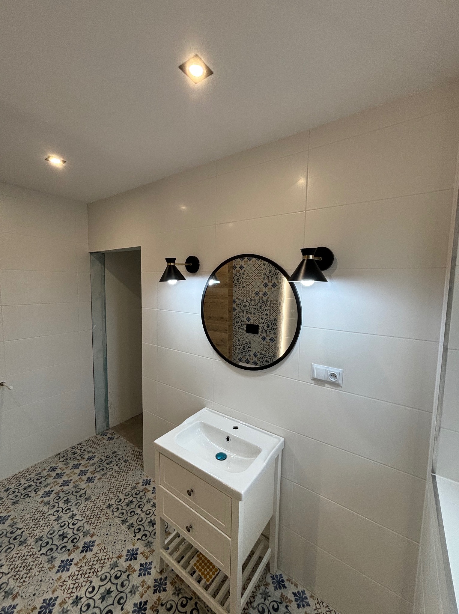 Wykończona łazienka w Szczekocinach: białe płytki na ścianach, wzorzysta podłoga, okrągłe lustro z oświetleniem i biała szafka pod umywalką. Nowoczesny design.