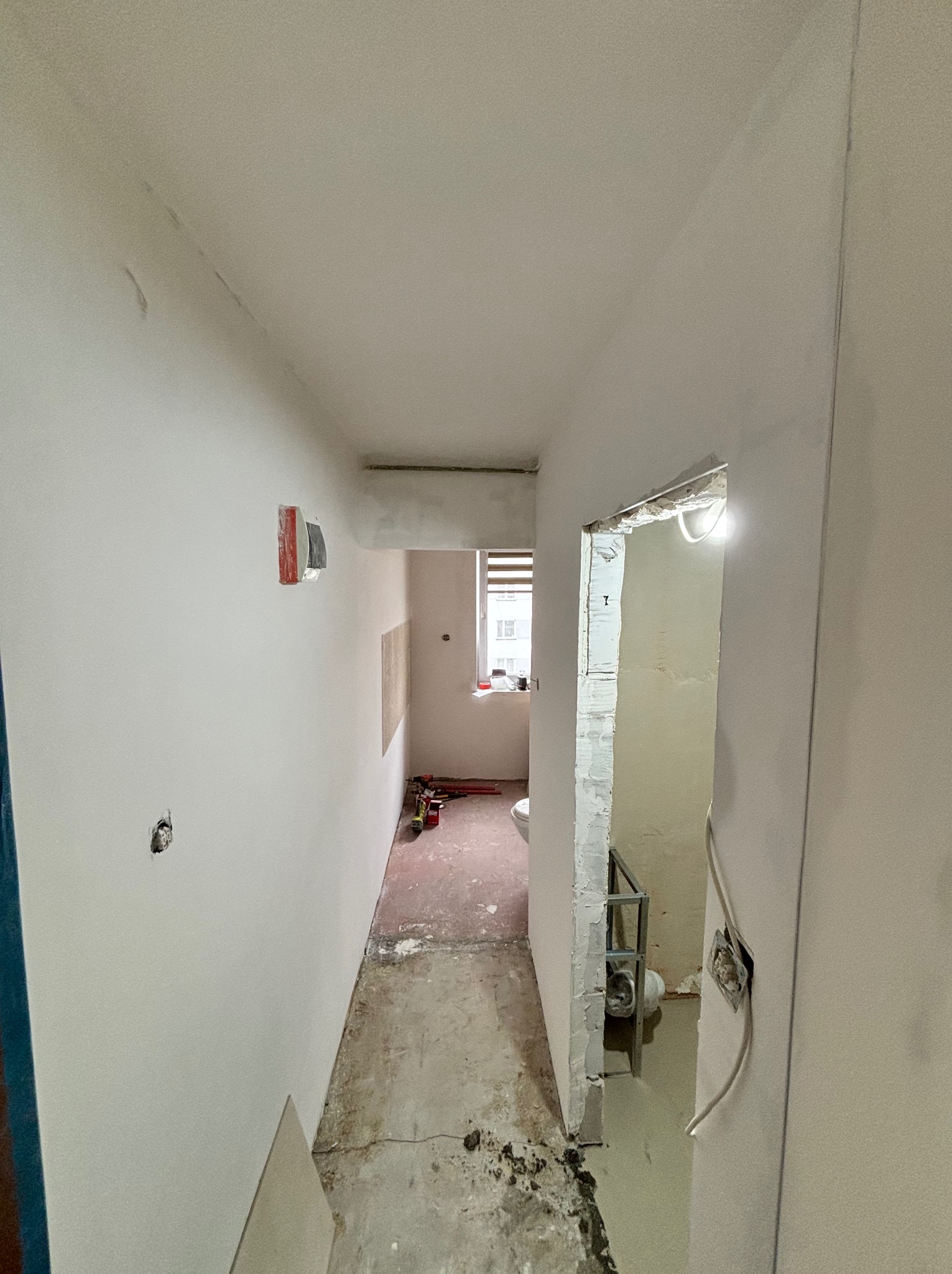 Korytarz w trakcie remontu: ściany po gładziowaniu, widoczne narzędzia, przygotowane otwory na gniazdka, surowa podłoga. Widok na łazienkę w trakcie wykończenia.
