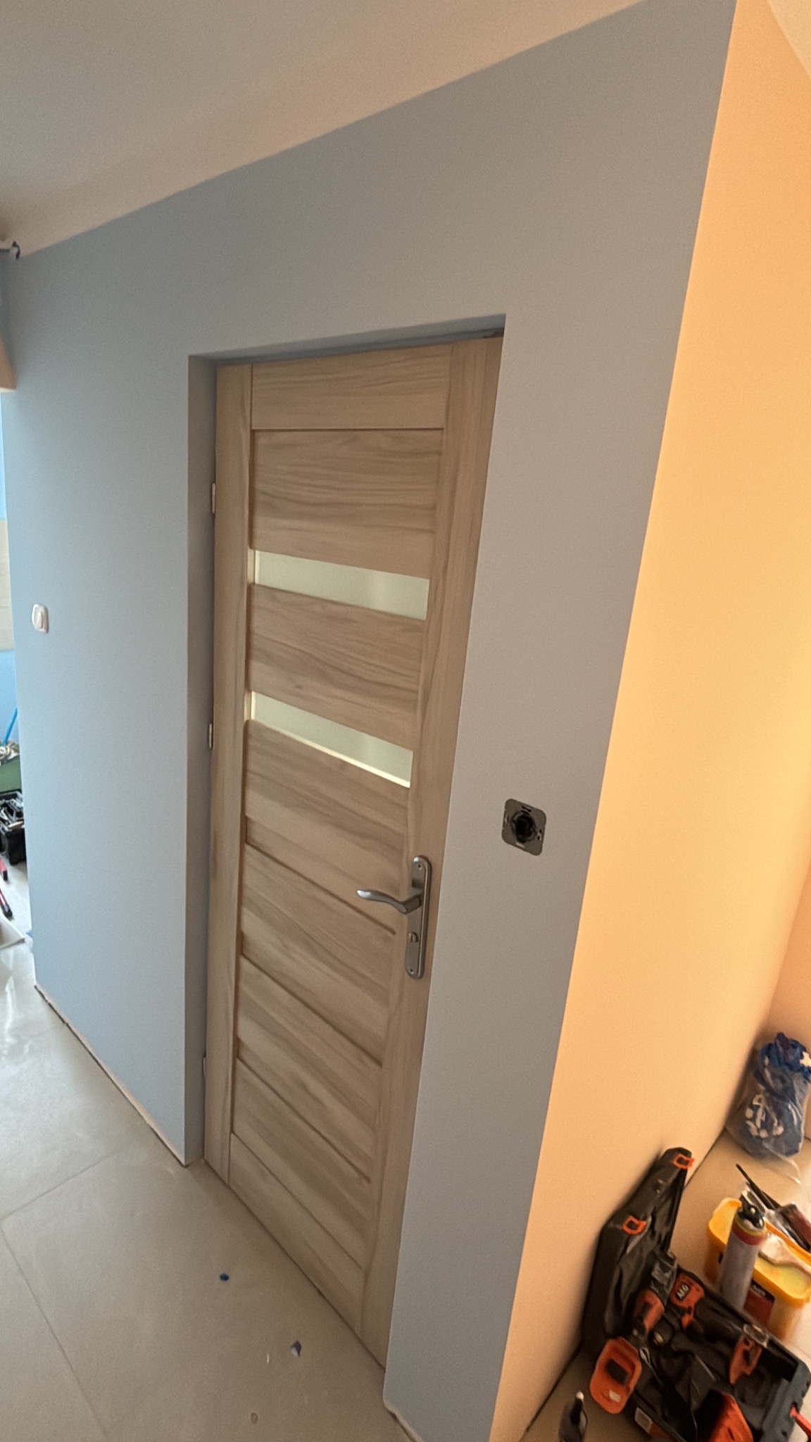 Jasne, drewniane drzwi z poziomymi przeszkleniami w świeżo pomalowanym na niebiesko pomieszczeniu. Widoczne narzędzia malarskie i gniazdko elektryczne.
