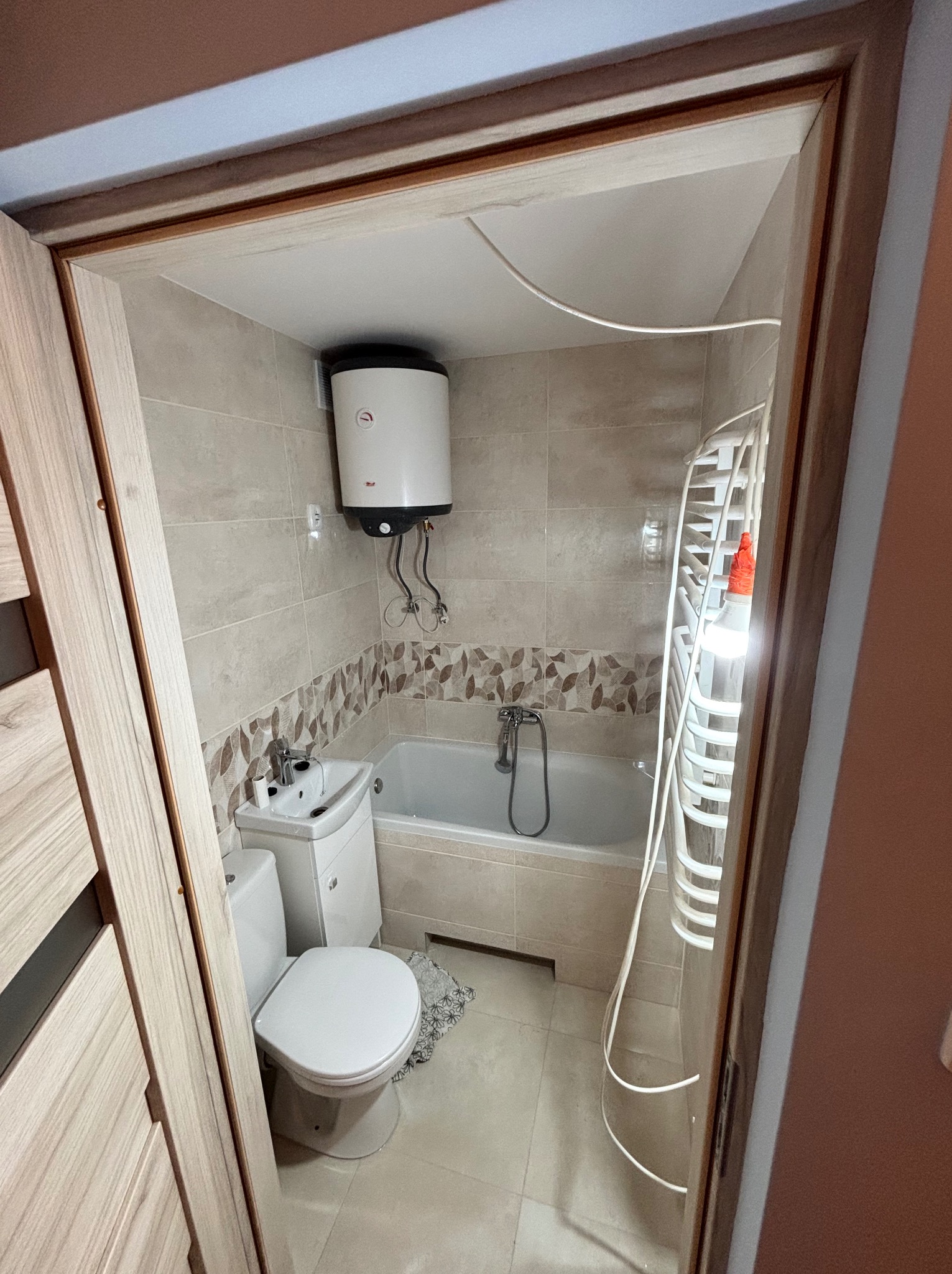 Jasna łazienka po remoncie: beżowe płytki, wanna, umywalka z szafką, toaleta i grzejnik drabinkowy. Widok zza drewnianej ramy drzwi.