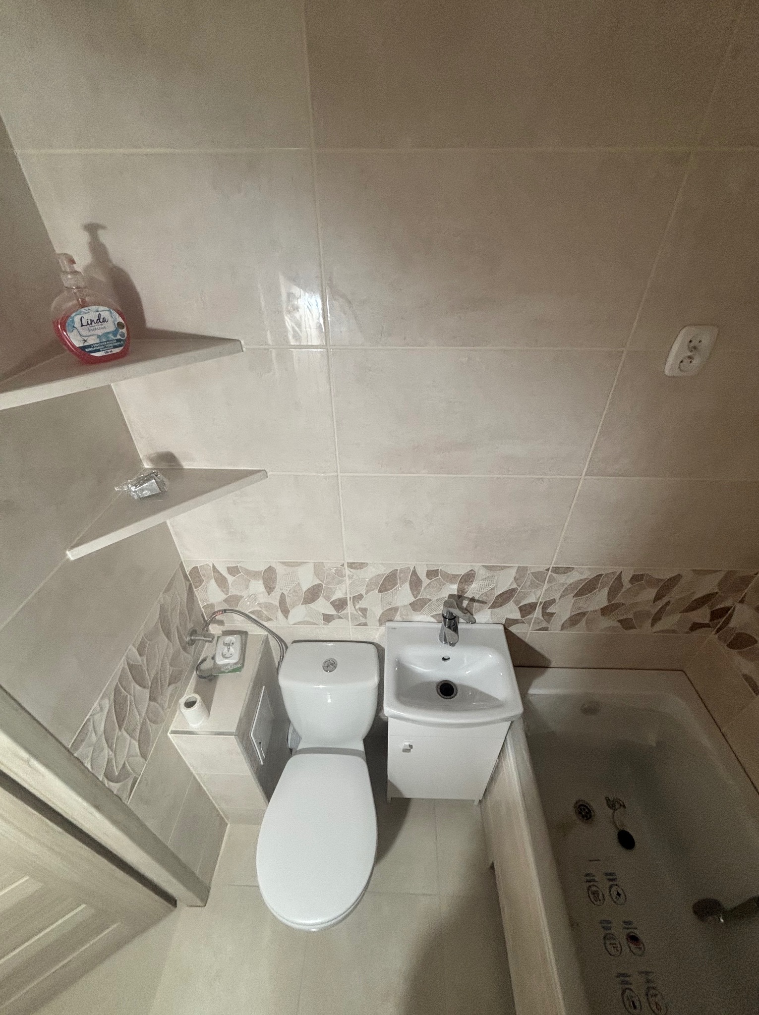 Jasna łazienka po remoncie: beżowe płytki, umywalka z szafką, toaleta i wanna z hydromasażem. Na półkach mydło i drobne akcesoria. Widok z góry.