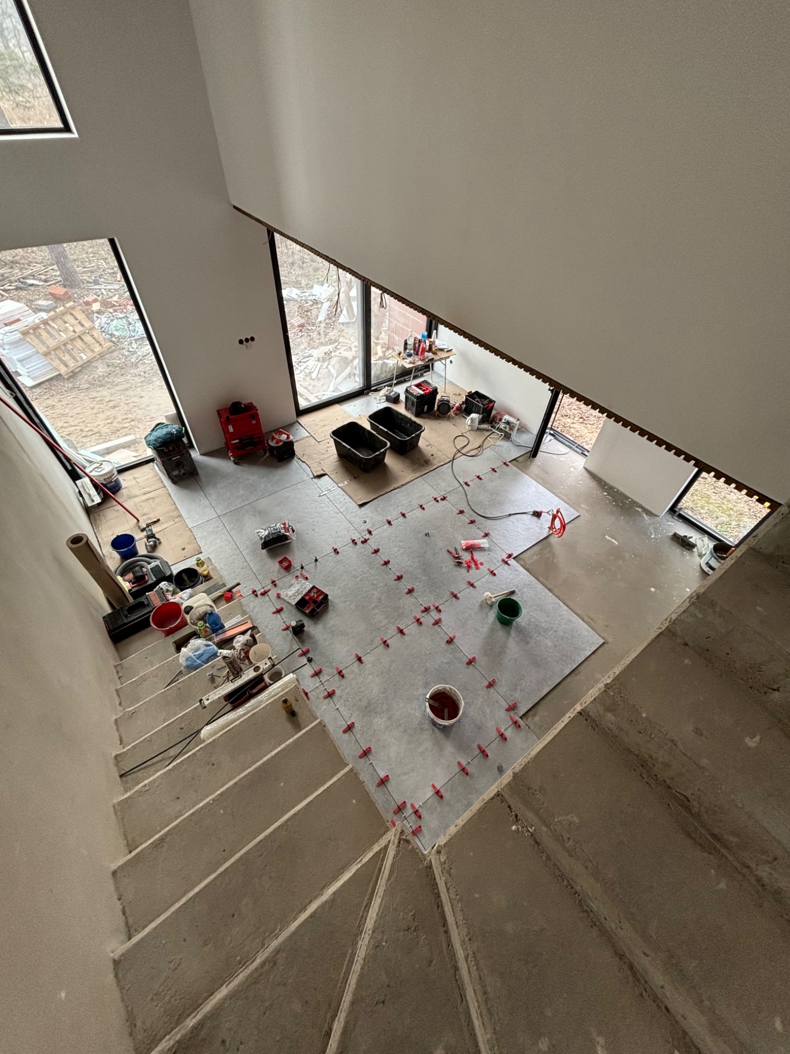 Układanie szarych płytek na podłodze w jasnym wnętrzu, widok z góry na schody i okna. Użyto czerwonych krzyżyków dystansowych. Część podłogi jeszcze nie wykończona.