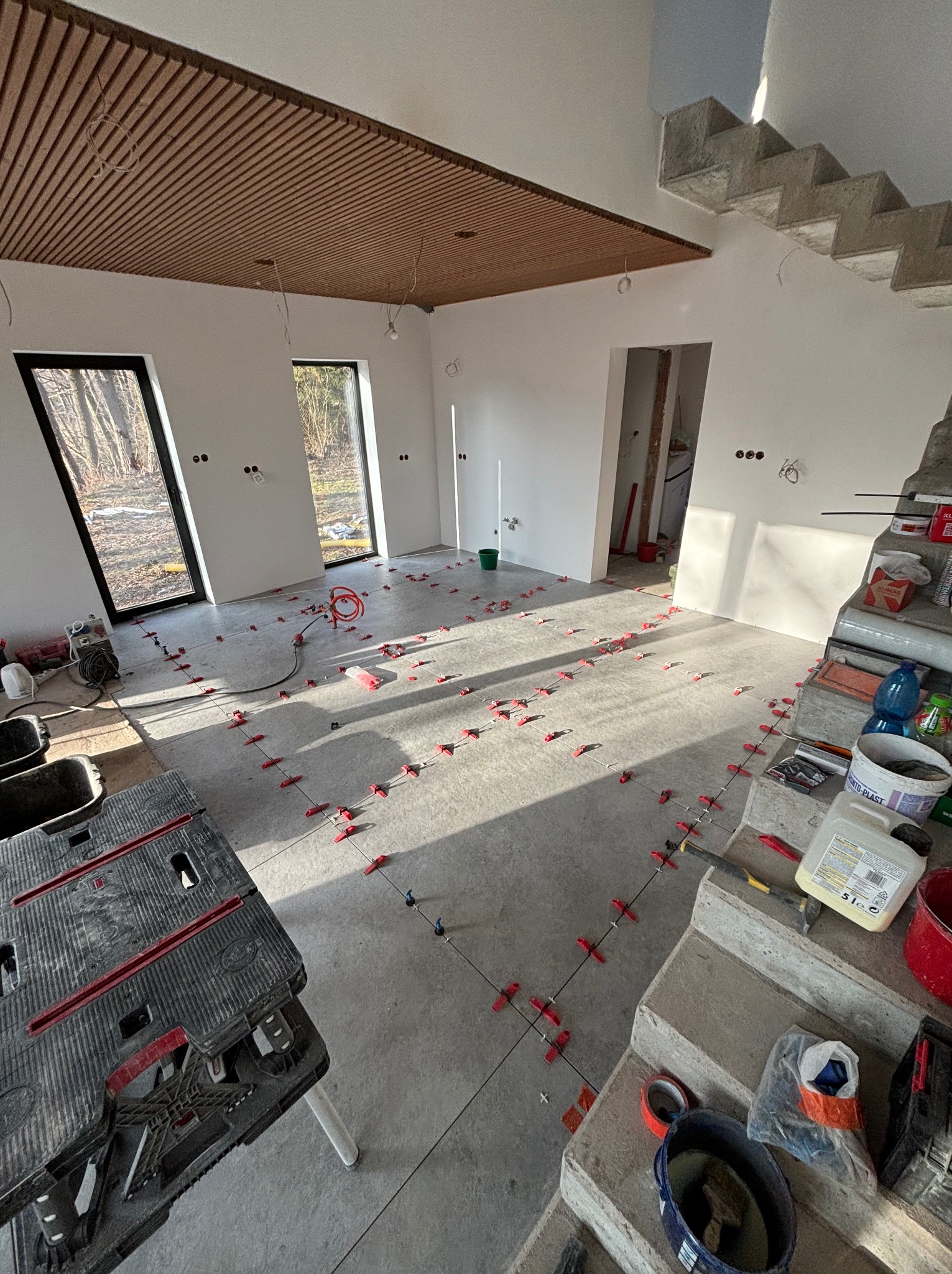 Układanie płytek podłogowych w jasnym wnętrzu z drewnianym sufitem lamelowym. Widoczne narzędzia glazurnicze i krzyżyki dystansowe.