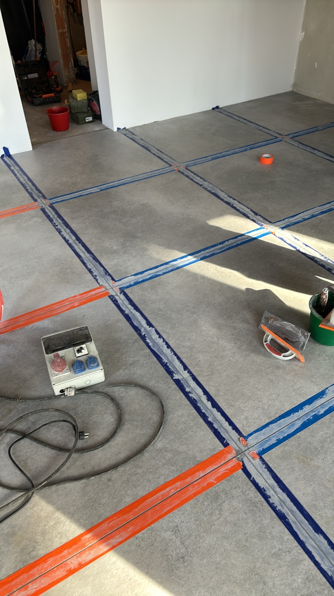 Przygotowanie podłoża pod wylewkę samopoziomującą z użyciem taśm dylatacyjnych w kolorze pomarańczowym i niebieskim. Widoczne narzędzia i puszka elektryczna.