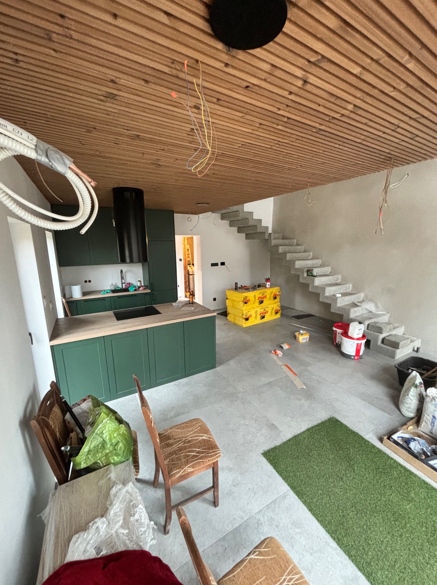Wnętrze w trakcie wykańczania: zielona zabudowa kuchenna, surowe betonowe schody, drewniany sufit lamelowy. Widoczne materiały budowlane i narzędzia.