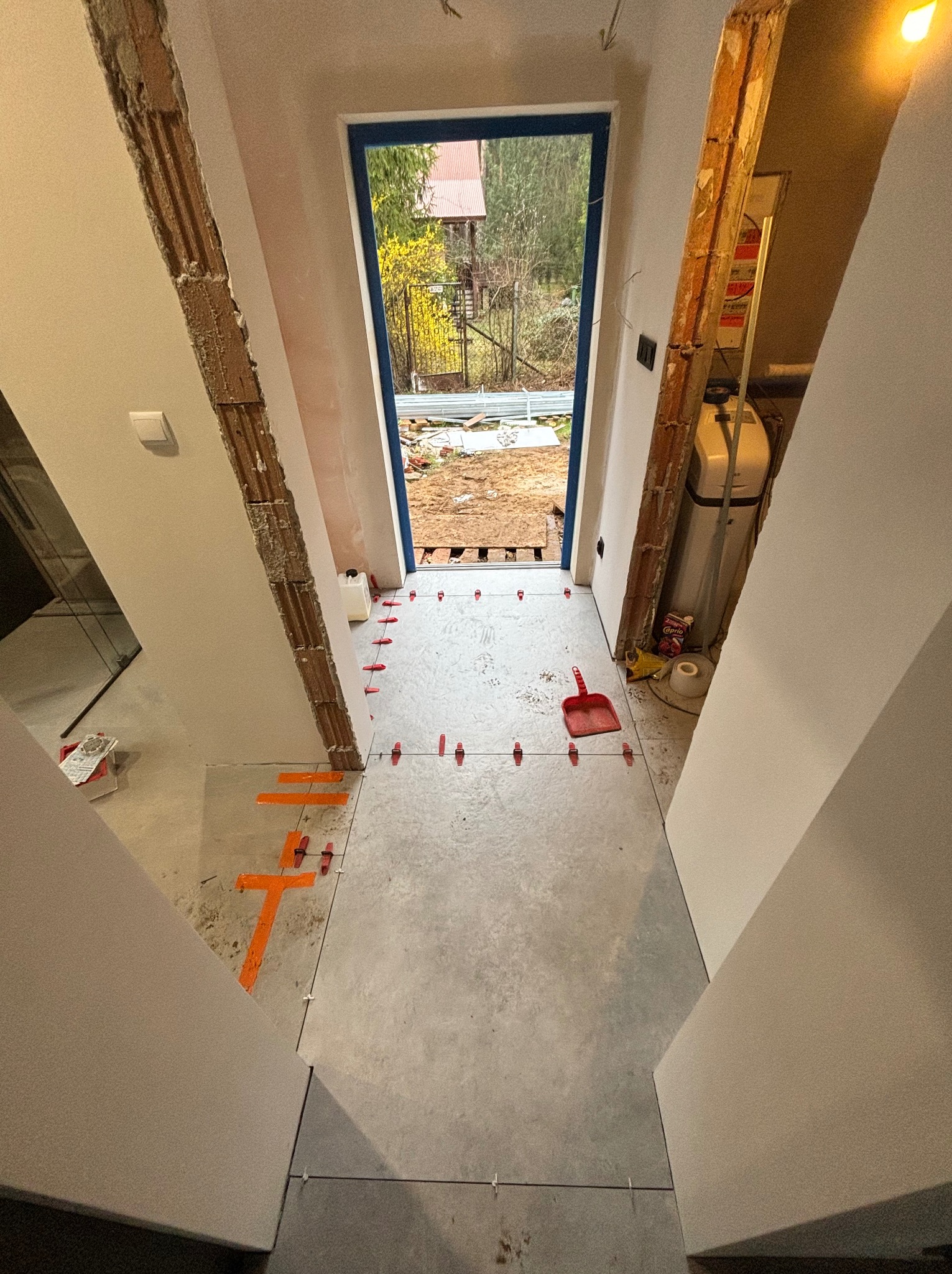 Układanie szarych płytek w korytarzu z widokiem na zewnątrz. Czerwone krzyżyki dystansowe i poziomica pomagają w precyzyjnym montażu. Widoczne przygotowane ściany.