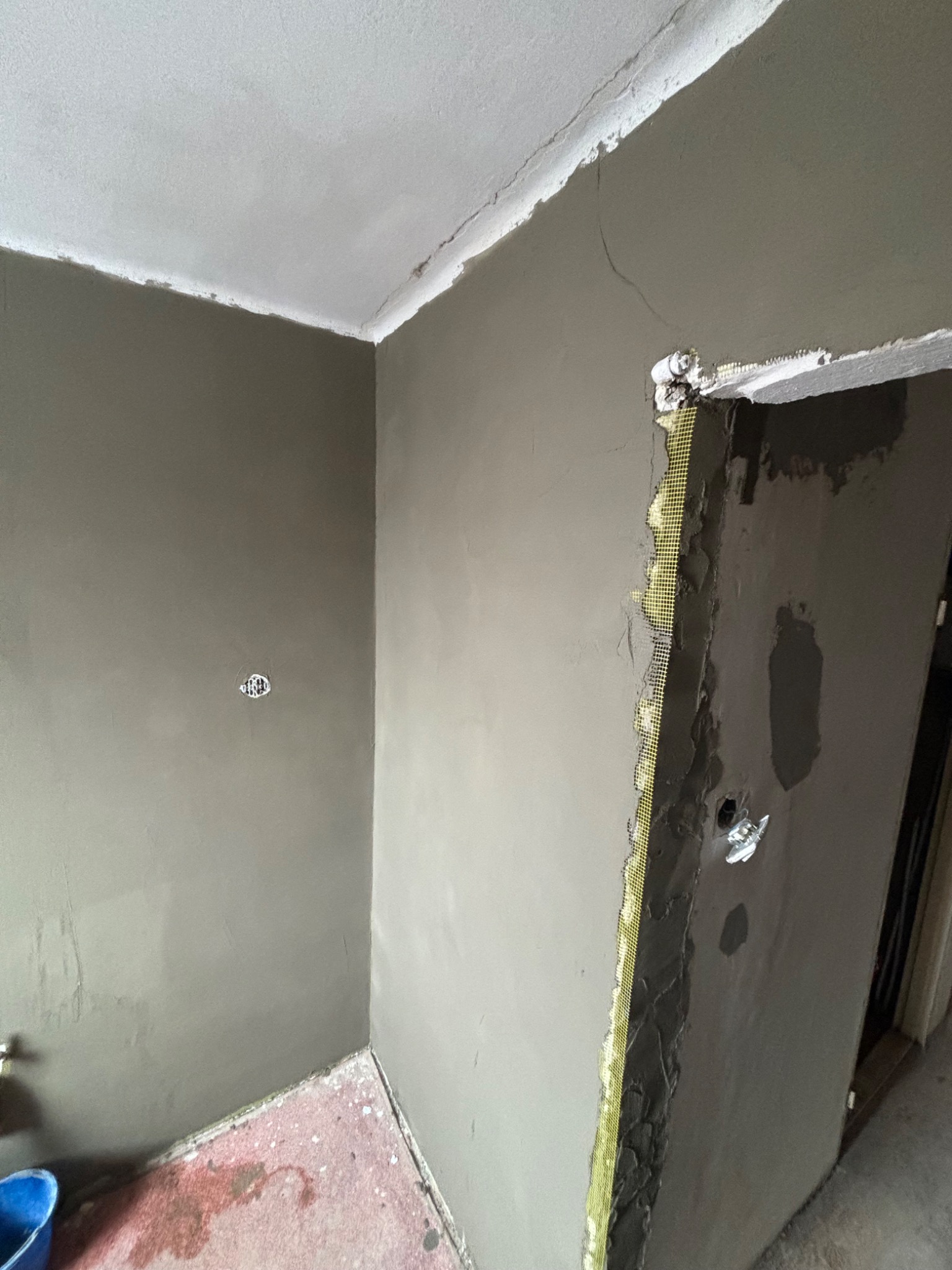 Ściana po nałożeniu szarej gładzi, widoczna siatka wzmacniająca przy ościeżnicy. Gniazdko elektryczne częściowo zakryte gładzią. Prace wykończeniowe w trakcie.