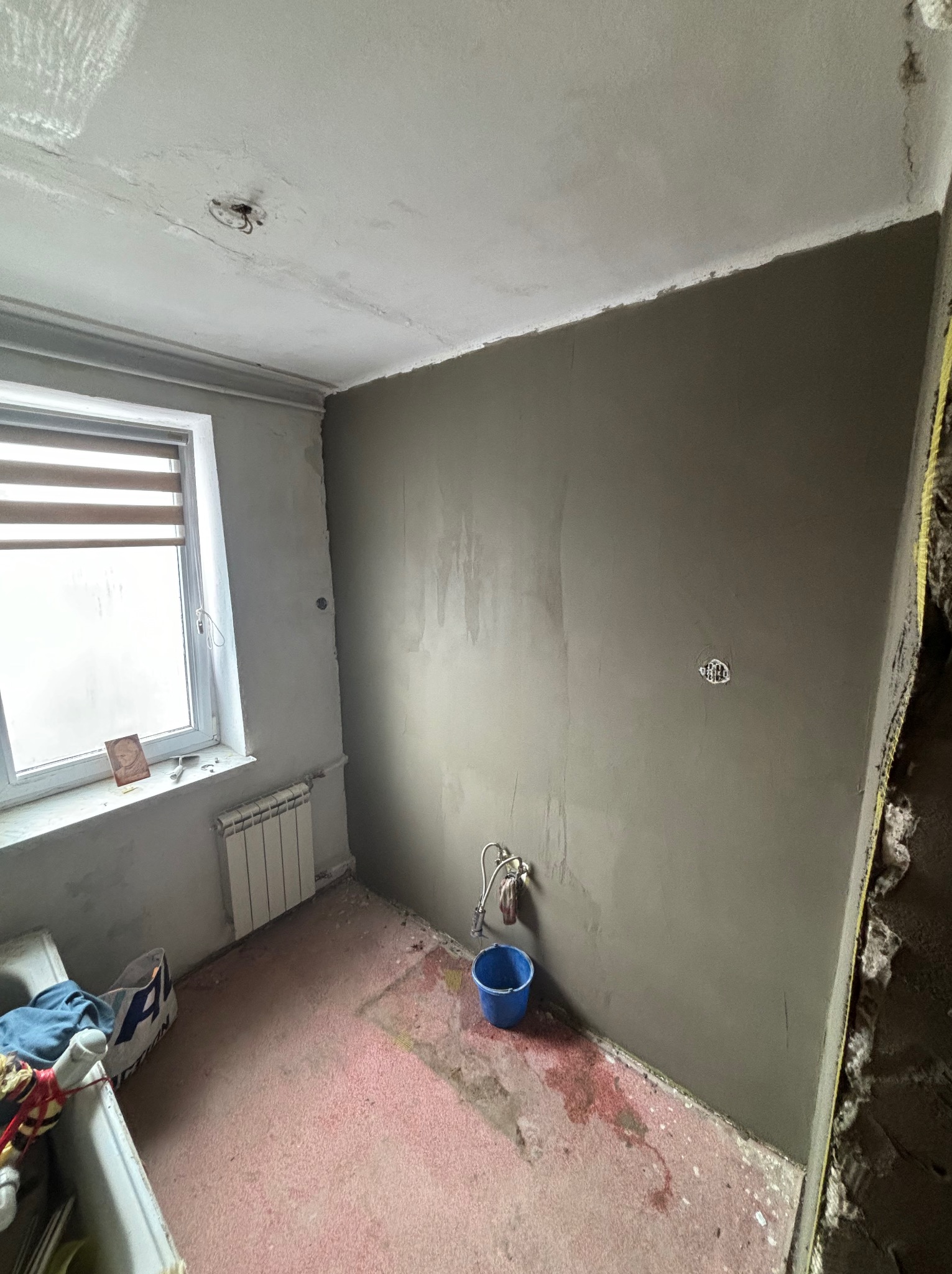 Pokój w trakcie remontu: ściana pokryta świeżą gładzią, widoczne instalacje, okno z roletami i grzejnik. Na podłodze ślady farby i wiadro.