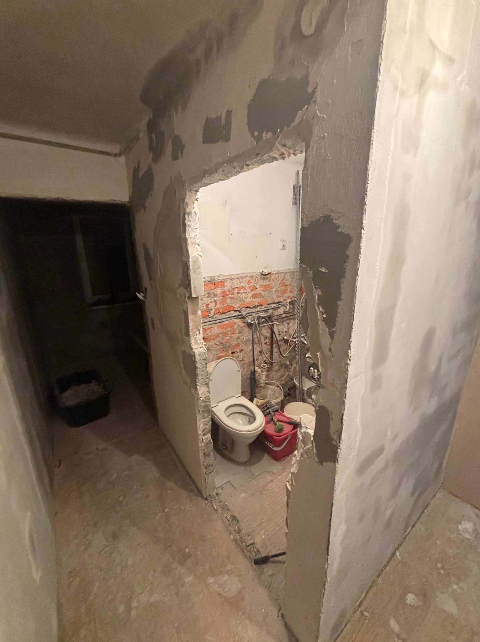 Łazienka w trakcie remontu: ściany z częściowo położoną gładzią, widoczna instalacja, toaleta. W tle wiadro z zaprawą. Ujęcie z perspektywy korytarza.