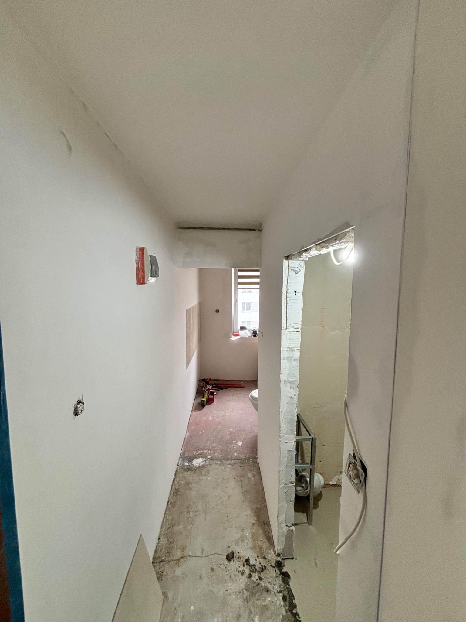 Wnętrze w trakcie remontu: białe ściany z widocznymi puszkami elektrycznymi, surowa podłoga i otwór drzwiowy do łazienki z widoczną instalacją.