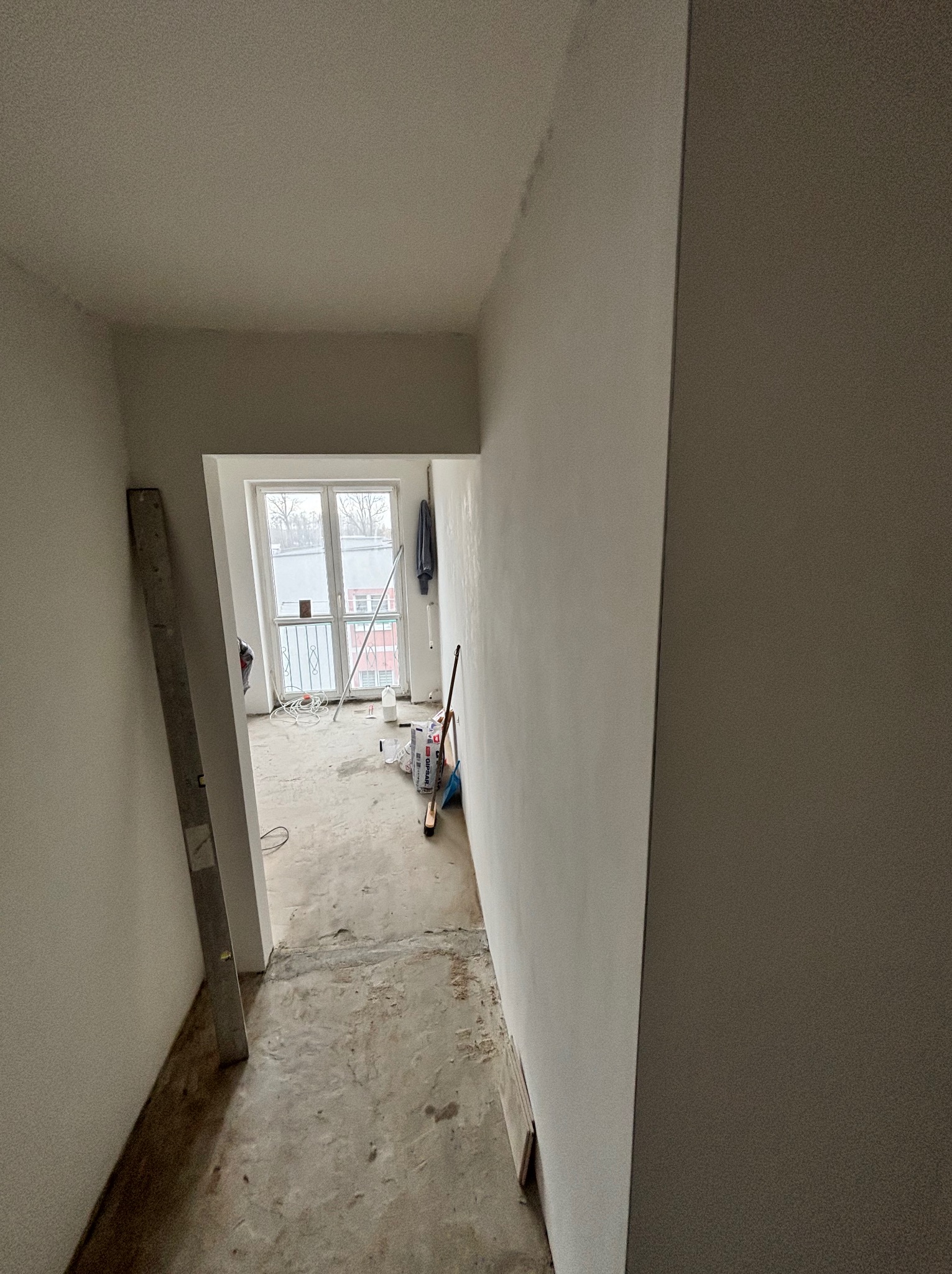 Wnętrze w trakcie remontu: świeżo pomalowane ściany na biało, widoczne narzędzia i materiały budowlane, okno z widokiem na zewnątrz. Minimalistyczny kadr.