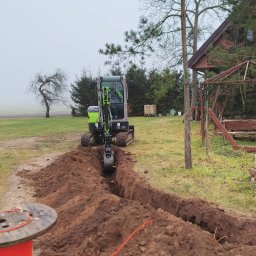 JW SMART SYSTEMS sp. z. o.o - Mini koparka z operatorem kopie wąski wykop w ziemi na zewnątrz, obok leży zwój pomarańczowego kabla. W tle dom i drzewa.