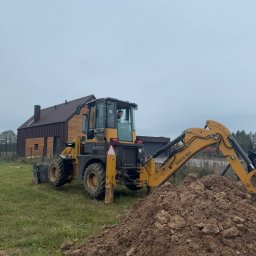 JW SMART SYSTEMS sp. z. o.o - Żółta koparko-ładowarka Günstig 918 na trawiastym terenie z pryzmą ziemi, w tle nowoczesny dom z brązową elewacją i szarym niebem. Operator w kabinie.