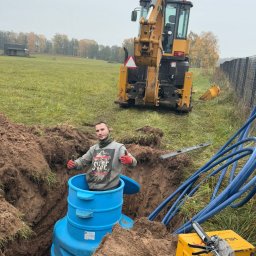 JW SMART SYSTEMS sp. z. o.o - Montaż niebieskiego zbiornika w wykopie, z pracownikiem dającym kciuki w górę, koparką w tle i niebieskimi rurami obok. Prace ziemne i instalacyjne w toku.