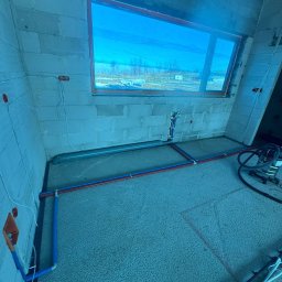 Instalacja wodna podłogowa w surowym wnętrzu z oknem. Rury w kolorze niebieskim i czerwonym ułożone na szarym podłożu. Widoczne przewody elektryczne na ścianach.