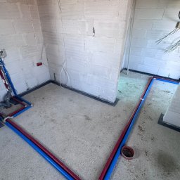 JW SMART SYSTEMS sp. z. o.o - Instalacja wodna w surowym wnętrzu: czerwone i niebieskie rury prowadzone po podłodze wzdłuż ścian z betonu komórkowego, widoczne złącza i fragment instalacji przy ścianie.