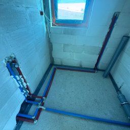 JW SMART SYSTEMS sp. z. o.o - Wnętrze w trakcie instalacji hydraulicznej: rury (czerwone, niebieskie) prowadzone wzdłuż ścian i podłogi, okno z widokiem na zewnątrz, ściany z białych bloczków.