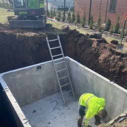 JW SMART SYSTEMS sp. z. o.o - Betonowy zbiornik w wykopie, drabina oparta o ścianę, pracownik w żółtej kamizelce, minikoparka w tle. Widok na budynek i ogrodzenie.